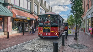 A red vintage-style trolley is driving along a cobblestone pedestrian street with brick buildings on either side. The building on the left has a 'Psychic Fair' sign, and there are several people walking nearby. A tree is planted to the right of the trolley, and the sky is partly cloudy.