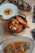 A variety of gourmet dishes are artistically arranged on a wooden table. A black cast iron skillet contains seafood including shrimp and mussels accompanied by a slice of toasted bread. Nearby, a white plate holds a creamy dish adorned with a small garnish. A rectangular black platter features meat or poultry pieces with sesame seeds and greens. At the bottom of the image, a stoneware dish presents sliced meat served in a flavorful sauce with garnishes of greens and red chili peppers. Two glasses of wine, one white and one red, complement the dining setup.