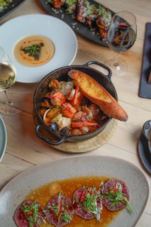 A variety of gourmet dishes are artistically arranged on a wooden table. A black cast iron skillet contains seafood including shrimp and mussels accompanied by a slice of toasted bread. Nearby, a white plate holds a creamy dish adorned with a small garnish. A rectangular black platter features meat or poultry pieces with sesame seeds and greens. At the bottom of the image, a stoneware dish presents sliced meat served in a flavorful sauce with garnishes of greens and red chili peppers. Two glasses of wine, one white and one red, complement the dining setup.
