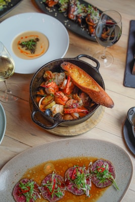 A variety of gourmet dishes are artistically arranged on a wooden table. A black cast iron skillet contains seafood including shrimp and mussels accompanied by a slice of toasted bread. Nearby, a white plate holds a creamy dish adorned with a small garnish. A rectangular black platter features meat or poultry pieces with sesame seeds and greens. At the bottom of the image, a stoneware dish presents sliced meat served in a flavorful sauce with garnishes of greens and red chili peppers. Two glasses of wine, one white and one red, complement the dining setup.