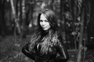 A woman with long hair stands confidently in a forest, wearing a leather jacket. The background is blurred, highlighting the texture of the surrounding trees and foliage.