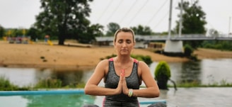 A person is meditating outdoors near a body of water and a bridge. They are seated on a blue yoga mat, wearing a gray and pink athletic outfit. The background includes a sandy area, trees, and a bridge with vertical supports.