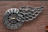 A wall-mounted metal sculpture features a circular design with a Chevrolet logo at its center and an intricate arrangement resembling a wing extending from it. The sculpture is made from shiny metallic components and is affixed to a red brick wall.