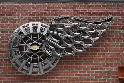 A wall-mounted metal sculpture features a circular design with a Chevrolet logo at its center and an intricate arrangement resembling a wing extending from it. The sculpture is made from shiny metallic components and is affixed to a red brick wall.