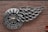 A wall-mounted metal sculpture features a circular design with a Chevrolet logo at its center and an intricate arrangement resembling a wing extending from it. The sculpture is made from shiny metallic components and is affixed to a red brick wall.