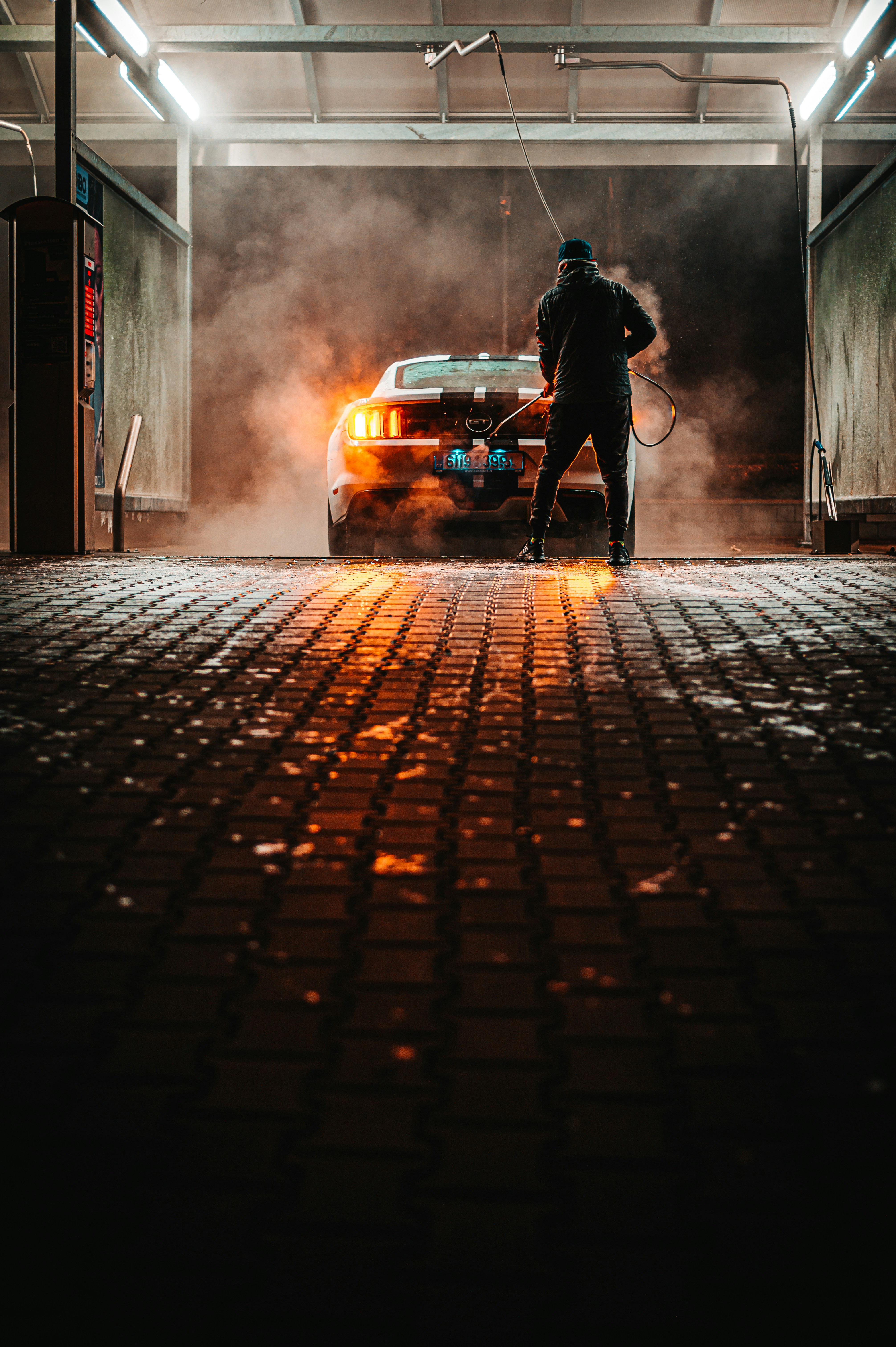 A person is washing a car at a dimly lit car wash. The car is parked under a roof with industrial lighting, creating a dramatic atmosphere with steam or smoke surrounding it. The pavement is wet, reflecting the orange glow from the car's rear lights.
