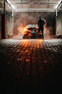 A person is washing a car at a dimly lit car wash. The car is parked under a roof with industrial lighting, creating a dramatic atmosphere with steam or smoke surrounding it. The pavement is wet, reflecting the orange glow from the car's rear lights.