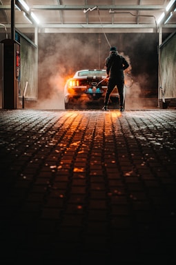 A person is washing a car at a dimly lit car wash. The car is parked under a roof with industrial lighting, creating a dramatic atmosphere with steam or smoke surrounding it. The pavement is wet, reflecting the orange glow from the car's rear lights.