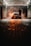 A person is washing a car at a dimly lit car wash. The car is parked under a roof with industrial lighting, creating a dramatic atmosphere with steam or smoke surrounding it. The pavement is wet, reflecting the orange glow from the car's rear lights.