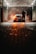 A person is washing a car at a dimly lit car wash. The car is parked under a roof with industrial lighting, creating a dramatic atmosphere with steam or smoke surrounding it. The pavement is wet, reflecting the orange glow from the car's rear lights.