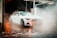 A sleek, white sports car in the midst of a car wash. The vehicle is surrounded by mist and steam, reflecting the light that creates a dramatic atmosphere. The setting appears to be at night, with wet, shining pavement beneath the car and an illuminated, slightly blurred poster or advertisement in the background.