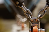 A close-up view of a metal chain with shiny, polished links connected to a metallic component featuring an orange turning mechanism. The background is blurred, creating a bokeh effect with circular light reflections.