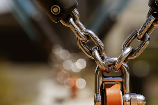 A close-up view of a metal chain with shiny, polished links connected to a metallic component featuring an orange turning mechanism. The background is blurred, creating a bokeh effect with circular light reflections.