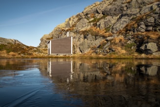 A modern, minimalist cabin is nestled against a rugged rocky hillside, surrounded by sparse vegetation. It stands beside a calm body of water that reflects the cabin and the earthy tones of the rocks. The landscape is serene, with clear blue skies above, emphasizing the tranquility of the natural setting.