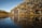 A modern, minimalist cabin is nestled against a rugged rocky hillside, surrounded by sparse vegetation. It stands beside a calm body of water that reflects the cabin and the earthy tones of the rocks. The landscape is serene, with clear blue skies above, emphasizing the tranquility of the natural setting.