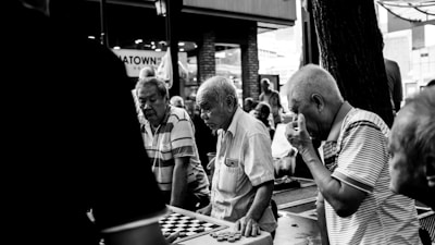 A group of elderly men is gathered around a table playing a board game in an outdoor setting. The scene captures their concentration and camaraderie. The setting appears to be a casual, possibly urban environment.