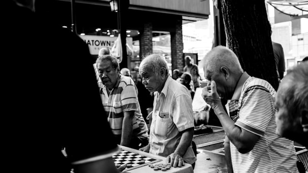 A group of elderly men is gathered around a table playing a board game in an outdoor setting. The scene captures their concentration and camaraderie. The setting appears to be a casual, possibly urban environment.