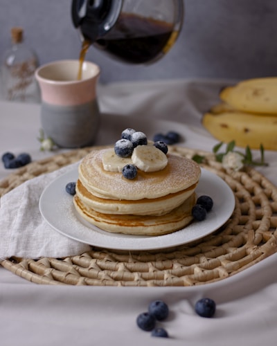 A white plate holds a stack of fluffy pancakes topped with banana slices and blueberries, sprinkled with powdered sugar. A woven placemat lies beneath, and fresh blueberries are scattered around. A ceramic mug is positioned nearby, being filled with freshly brewed coffee from a glass carafe. The background features a soft-focus display of bananas and a small bottle, creating a cozy breakfast setting.