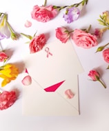 An arrangement of various flowers, predominantly pink roses, surrounds an envelope with a pink awareness ribbon on it. The composition is placed on a white surface, with rose petals scattered around.