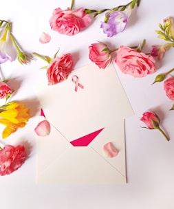 An arrangement of various flowers, predominantly pink roses, surrounds an envelope with a pink awareness ribbon on it. The composition is placed on a white surface, with rose petals scattered around.