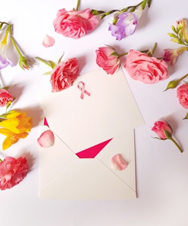 An arrangement of various flowers, predominantly pink roses, surrounds an envelope with a pink awareness ribbon on it. The composition is placed on a white surface, with rose petals scattered around.