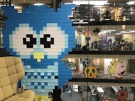 A large blue owl is created using sticky notes on a glass wall in an office setting. Other pixelated characters are seen on various floors of the open office space, all made from colorful sticky notes. The office has a modern and creative ambiance.