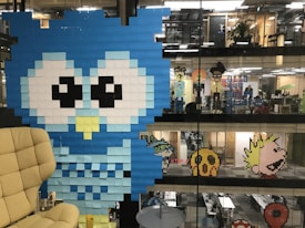 A large blue owl is created using sticky notes on a glass wall in an office setting. Other pixelated characters are seen on various floors of the open office space, all made from colorful sticky notes. The office has a modern and creative ambiance.