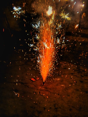 A bright burst of colorful sparks erupts from a firecracker on the ground, creating a spray of orange, blue, and white light against a dark background. Smaller star-like sparks are visible in the surrounding area.
