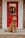 A fluffy dog with curly fur is sitting on a front porch in front of a vibrant red door adorned with a festive wreath. The house number, 282, is visible beside the door, and the porch is made of brick.