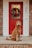 A fluffy dog with curly fur is sitting on a front porch in front of a vibrant red door adorned with a festive wreath. The house number, 282, is visible beside the door, and the porch is made of brick.