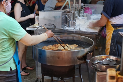 A bustling street food scene featuring a person frying food in a large metal wok. The cook uses long wooden chopsticks to manage the food, which appears to be golden brown and crispy. The surrounding area has a variety of kitchen tools and ingredients, including flour, utensils, and metal containers. Other people are visible in the background, suggesting a lively and busy environment.