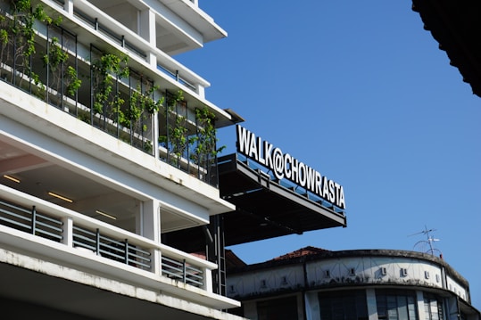 A modern building with plants growing along its vertical rails contrasts against a clear blue sky. The sign 'WALK@CHOWRASTA' is prominently displayed, attached to a structure slightly above the foreground. Another building with a more traditional design and an antenna is visible in the background.