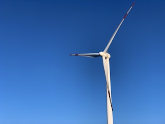 A tall wind turbine with three blades set against a clear blue sky. The blades have red markings at the tips for visibility.