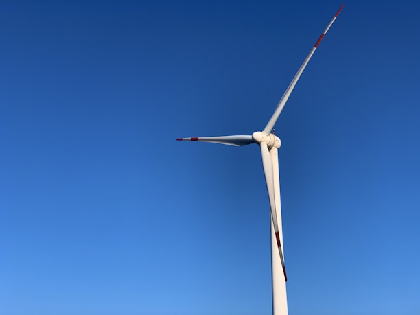 A tall wind turbine with three blades set against a clear blue sky. The blades have red markings at the tips for visibility.