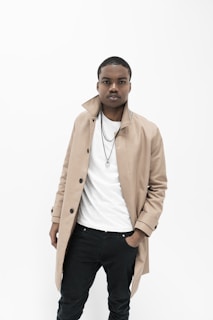 A person stands confidently against a white background, wearing a beige trench coat over a white shirt, paired with black pants. A couple of necklaces add a subtle touch of style.