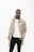 A person stands confidently against a white background, wearing a beige trench coat over a white shirt, paired with black pants. A couple of necklaces add a subtle touch of style.