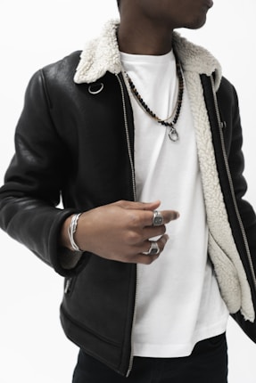 A person is wearing a black leather jacket with a white inner lining over a plain white t-shirt. They have several accessories, including silver rings and a bracelet, alongside a beaded necklace with a pendant.
