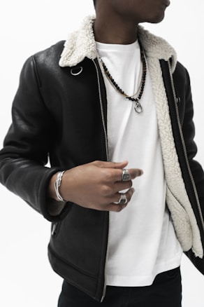 A person is wearing a black leather jacket with a white inner lining over a plain white t-shirt. They have several accessories, including silver rings and a bracelet, alongside a beaded necklace with a pendant.