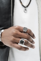 A close-up of a person's hand showcasing two large, ornate rings. The hand is highlighted against a blurred background, with a black leather jacket and white shirt. The rings have intricate designs, one resembling a watch face and the other with a rectangular pattern. A chain necklace is slightly visible above.