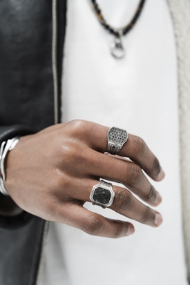 A close-up of a person's hand showcasing two large, ornate rings. The hand is highlighted against a blurred background, with a black leather jacket and white shirt. The rings have intricate designs, one resembling a watch face and the other with a rectangular pattern. A chain necklace is slightly visible above.