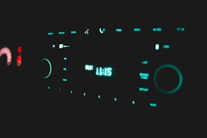 A close-up of a car stereo control panel illuminated by a soft teal light. The center display shows the time as 11:15 with the word 'AUX' indicating the input source. Surrounding the display are various buttons and knobs for controlling the audio system, including options for AM, FM, and disc.