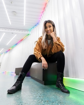 A smiling woman with long hair sits on a bench in a modern, brightly lit hallway. She is wearing a brown jacket, black leggings, and black boots. The hallway features white walls and ceiling with a colorful LED strip running along the edge.