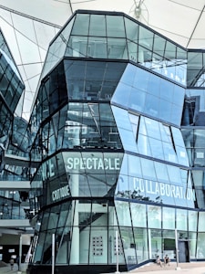 A modern architectural structure with angular glass facades reflecting the surroundings. Words like 'Spectacle' and 'Collaboration' are visible on the building's surface. The interior is partially visible, showing framed pictures on the walls and a few people near the entrance.