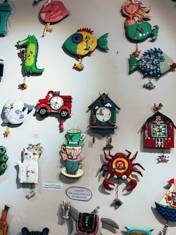 A collection of colorful, whimsical wall clocks and decorations featuring playful designs such as a cat, fish, crab, horse, teacups, and a red barn. Each piece is crafted with bold colors and imaginative shapes, creating a lively and artistic display.