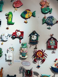 A collection of colorful, whimsical wall clocks and decorations featuring playful designs such as a cat, fish, crab, horse, teacups, and a red barn. Each piece is crafted with bold colors and imaginative shapes, creating a lively and artistic display.