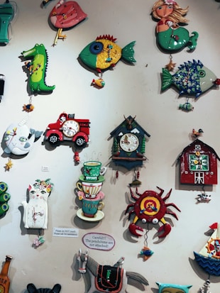 A collection of colorful, whimsical wall clocks and decorations featuring playful designs such as a cat, fish, crab, horse, teacups, and a red barn. Each piece is crafted with bold colors and imaginative shapes, creating a lively and artistic display.