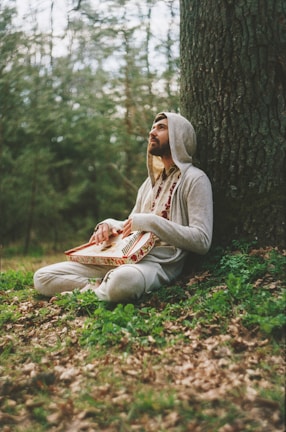 Rahu Dev in a quiet forest, sitting cross-legged with a harmonium, sunlight filtering through the trees.