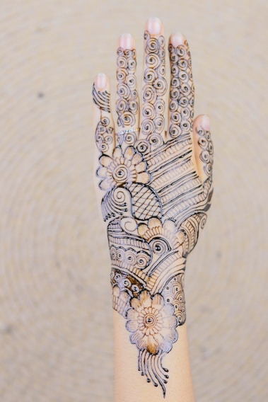 A hand is decorated with intricate henna designs featuring floral patterns, swirls, and lines. The artwork covers the entire back of the hand, extending to the fingers.