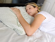 A person is peacefully sleeping on a bed, resting their head on a pillow with a relaxed expression. The white bedding and ambient lighting contribute to a calm and serene atmosphere.