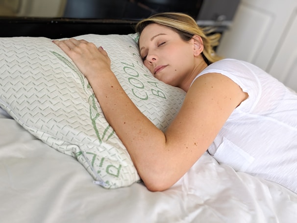 A person is peacefully sleeping on a bed, resting their head on a pillow with a relaxed expression. The white bedding and ambient lighting contribute to a calm and serene atmosphere.